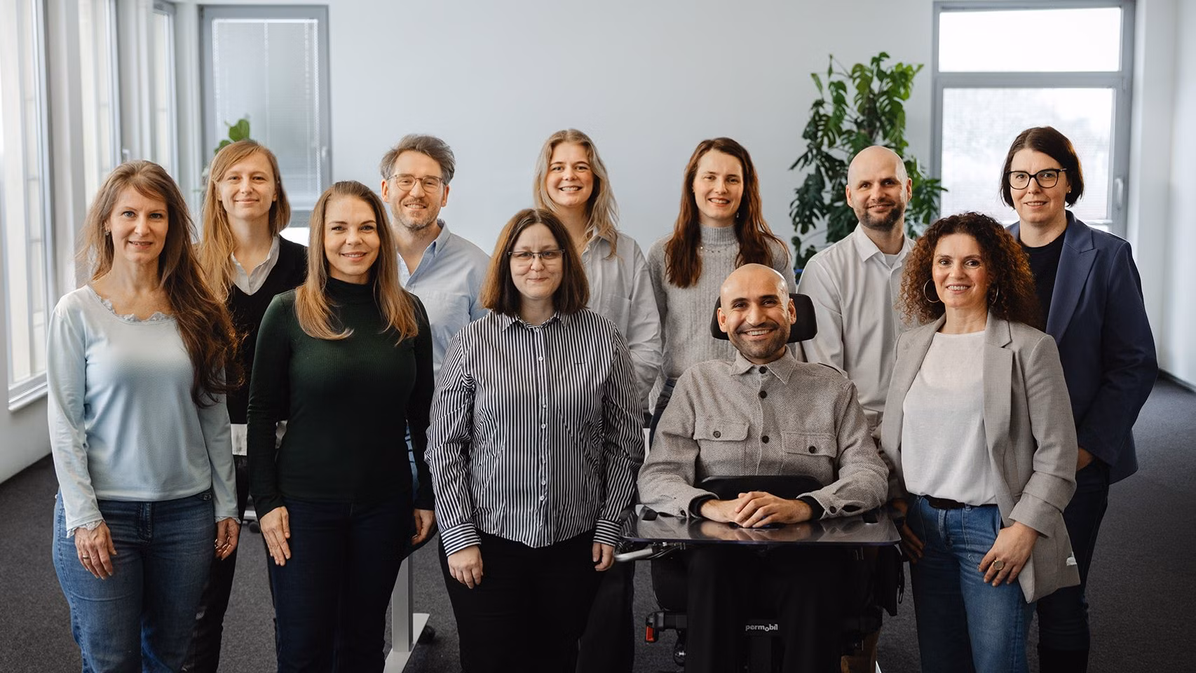 Team aus Mitarbeitenden steht gemeinsam in einem Büro, eine Person sitzt im Rollstuhl