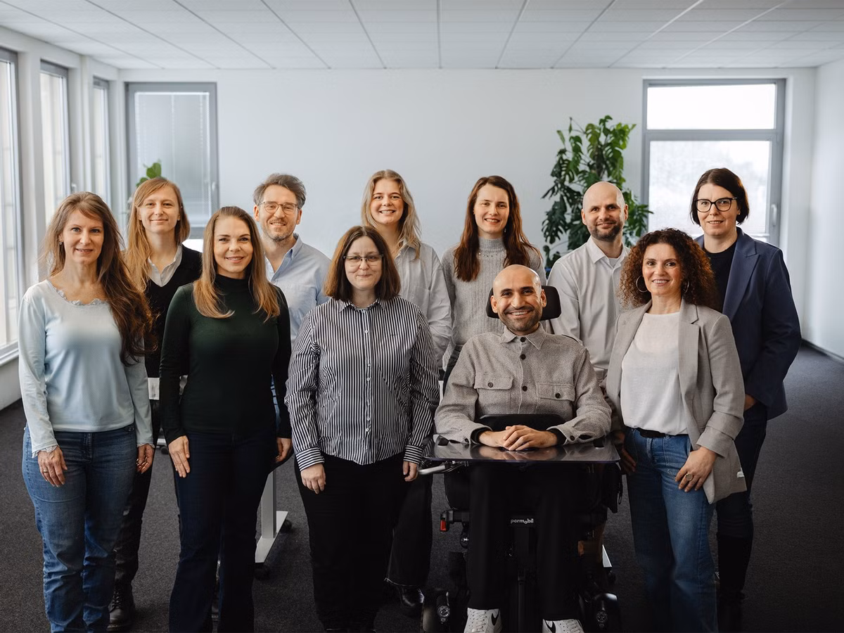 Team aus Mitarbeitenden steht gemeinsam in einem Büro, eine Person sitzt im Rollstuhl